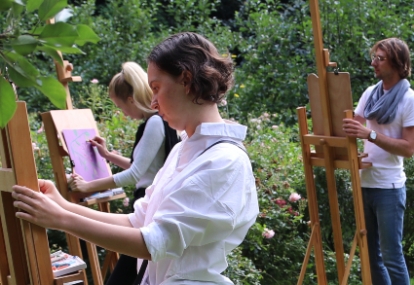 Trójka uczestników muzealnych warsztatów stoi w zielonym ogrodzie i maluje na płótnach rozstawionych na drewnianych sztalugach. 